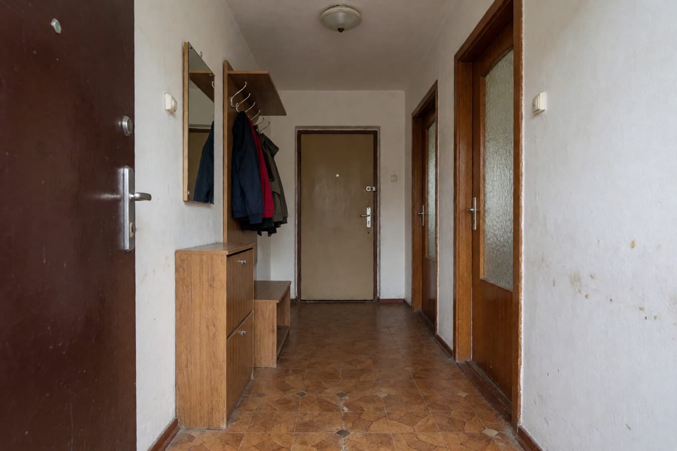 Hol de apartament vechi cu linoleum maro tocit, pantofar din lemn, cuier cu geci și uși interioare cu geam