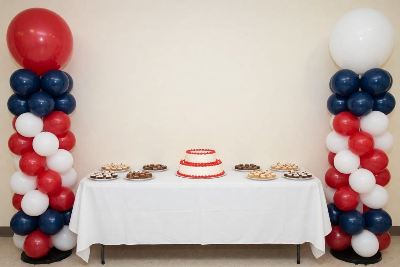 Dalawang balloon column na pula, puti at navy sa magkabilang dulo ng mahabang mesa na may two-tier na cake at mga matamis