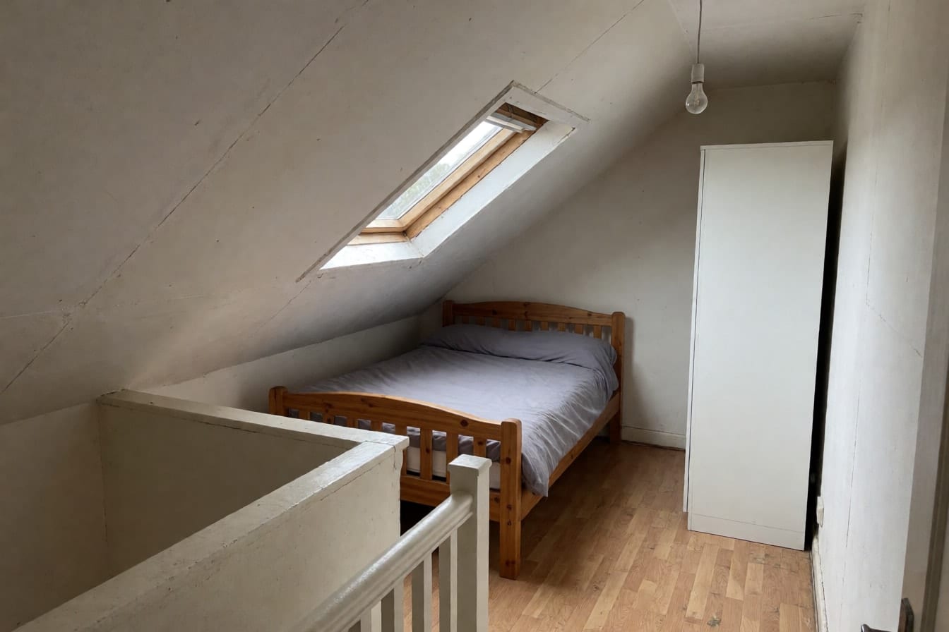 Kahles Dachgeschoss-Schlafzimmer mit weißer Dachschräge, Dachfenster, Kiefernbett unter der Schräge und nackter Glühbirne
