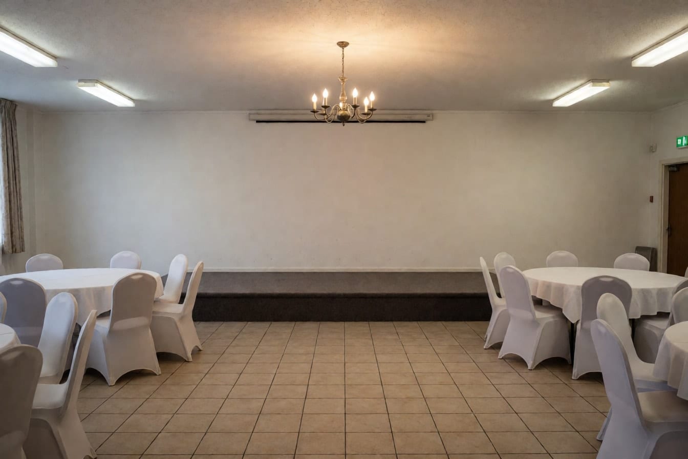 Bakanteng function hall na may mababang stage na may carpet sa harap ng puting dingding, bilog na mesa na may puting tela at upuang may cover