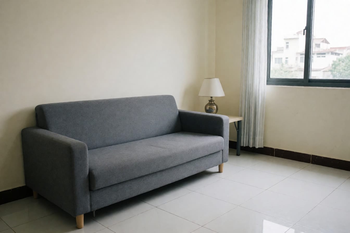 Góc phòng khách căn hộ với sofa nỉ xám sát tường kem, sàn gạch men trắng, bàn nhỏ có đèn và cửa sổ rèm trắng mỏng