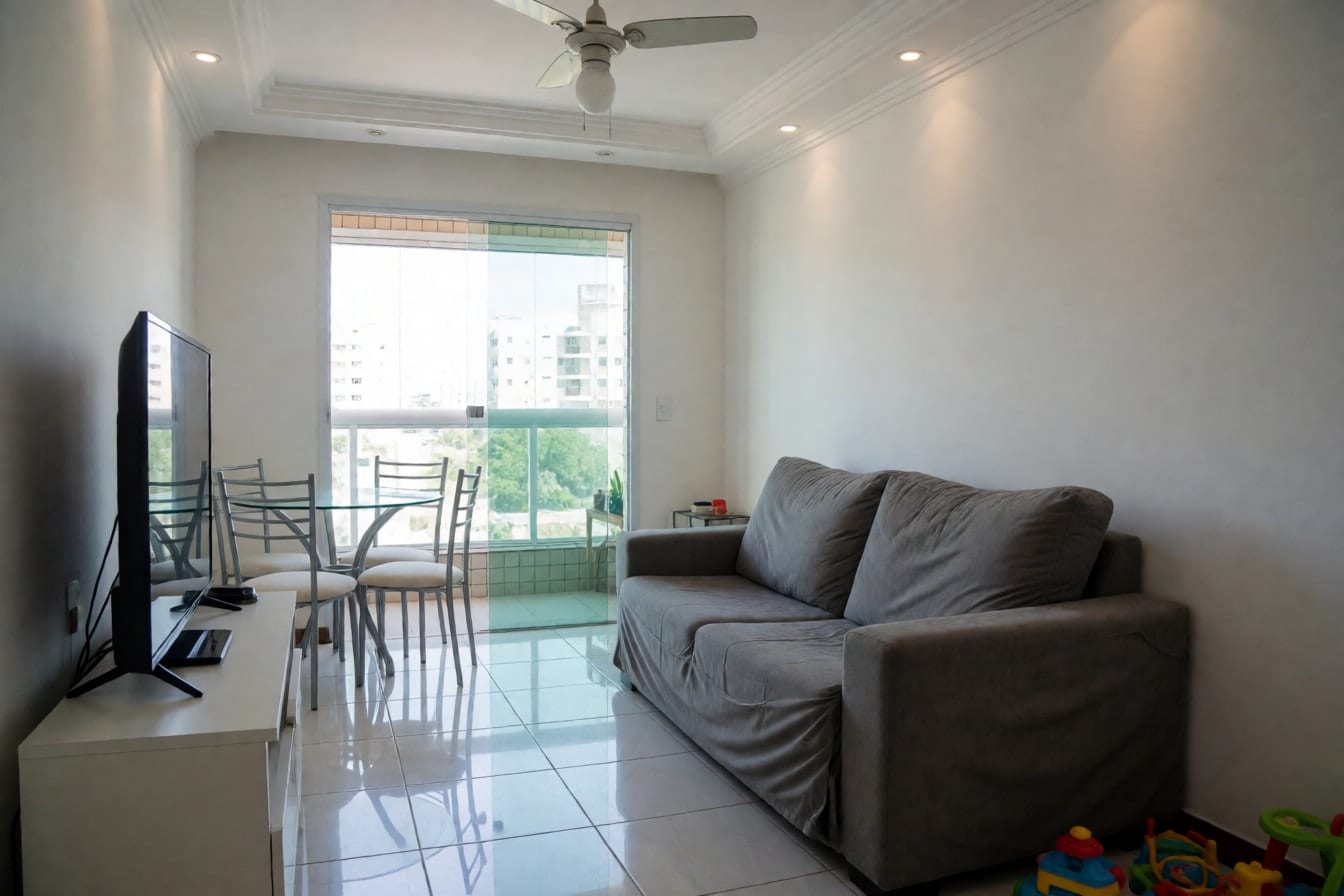 Sala de apartamento com sofá retrátil cinza, TV sobre rack branco, mesa redonda de vidro, piso de porcelanato branco e porta de vidro para a varanda