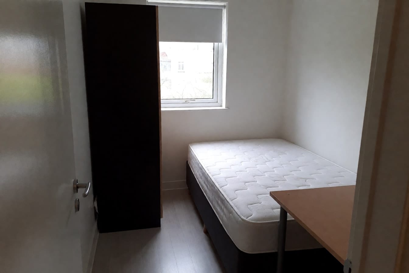 Kleines Schlafzimmer mit Doppelbett in der Raummitte, dunklem Kleiderschrank am Fenster, Schreibtisch am Fußende und schmalem Durchgang