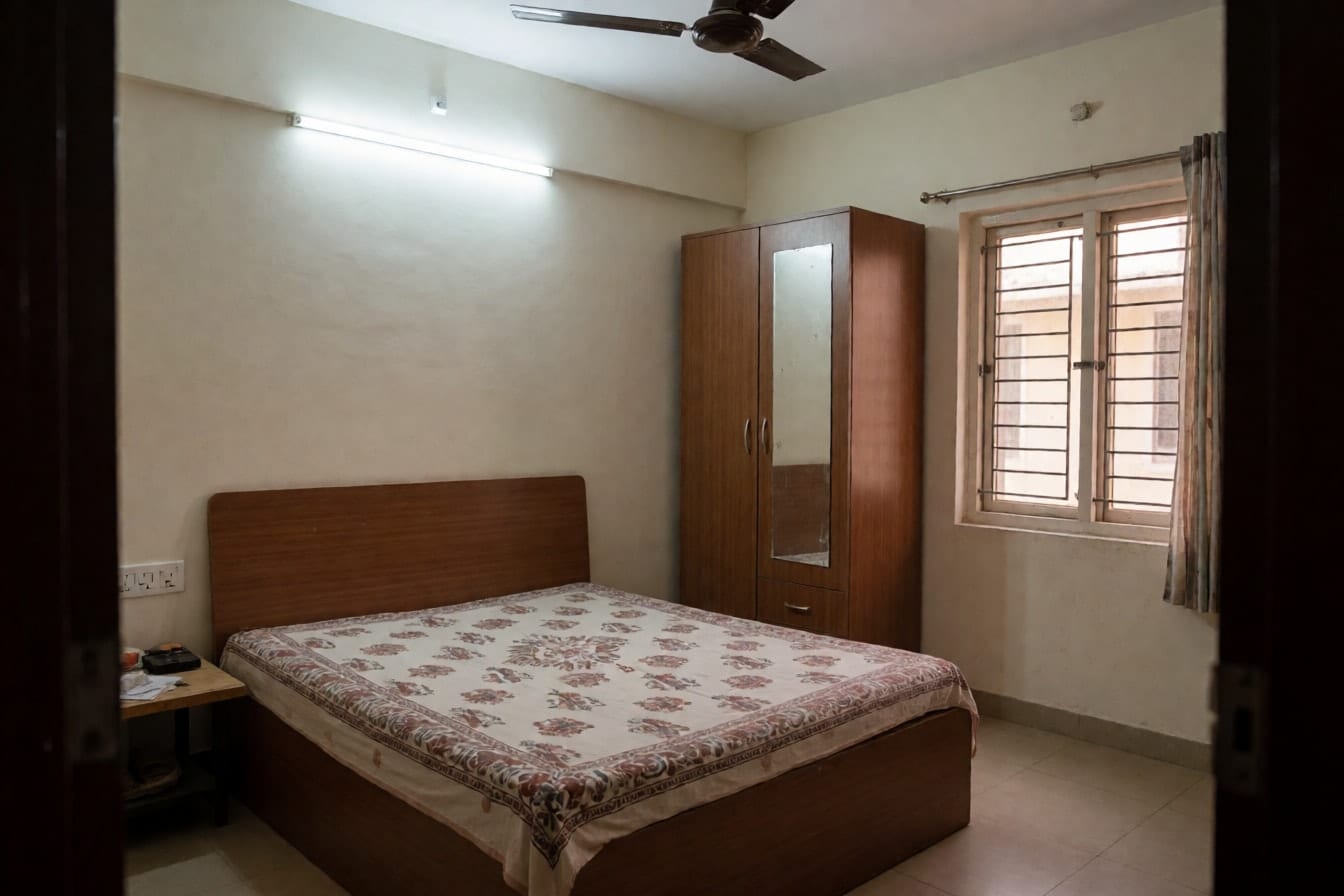 फ़्लैट का पुराना बेडरूम: डबल बेड पर छपी चादर, शीशे वाली भूरी लैमिनेट अलमारी, सफ़ेद दीवारें और ट्यूबलाइट