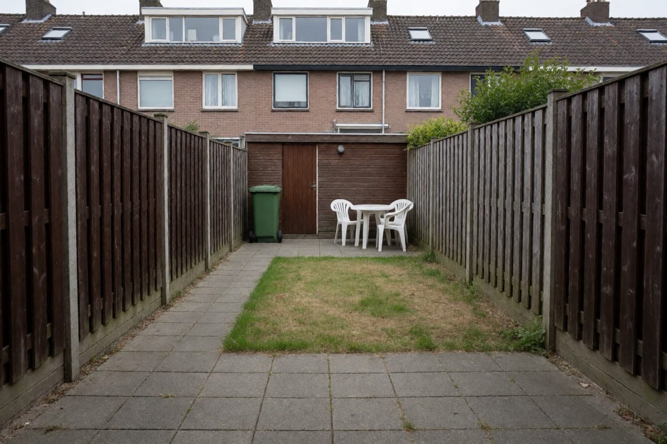 Smalle, diepe achtertuin van een rijtjeshuis met grijze betontegels, een klein stukje gras, hoge bruine schuttingen, een houten schuur, een groene kliko en een witte plastic tuinset