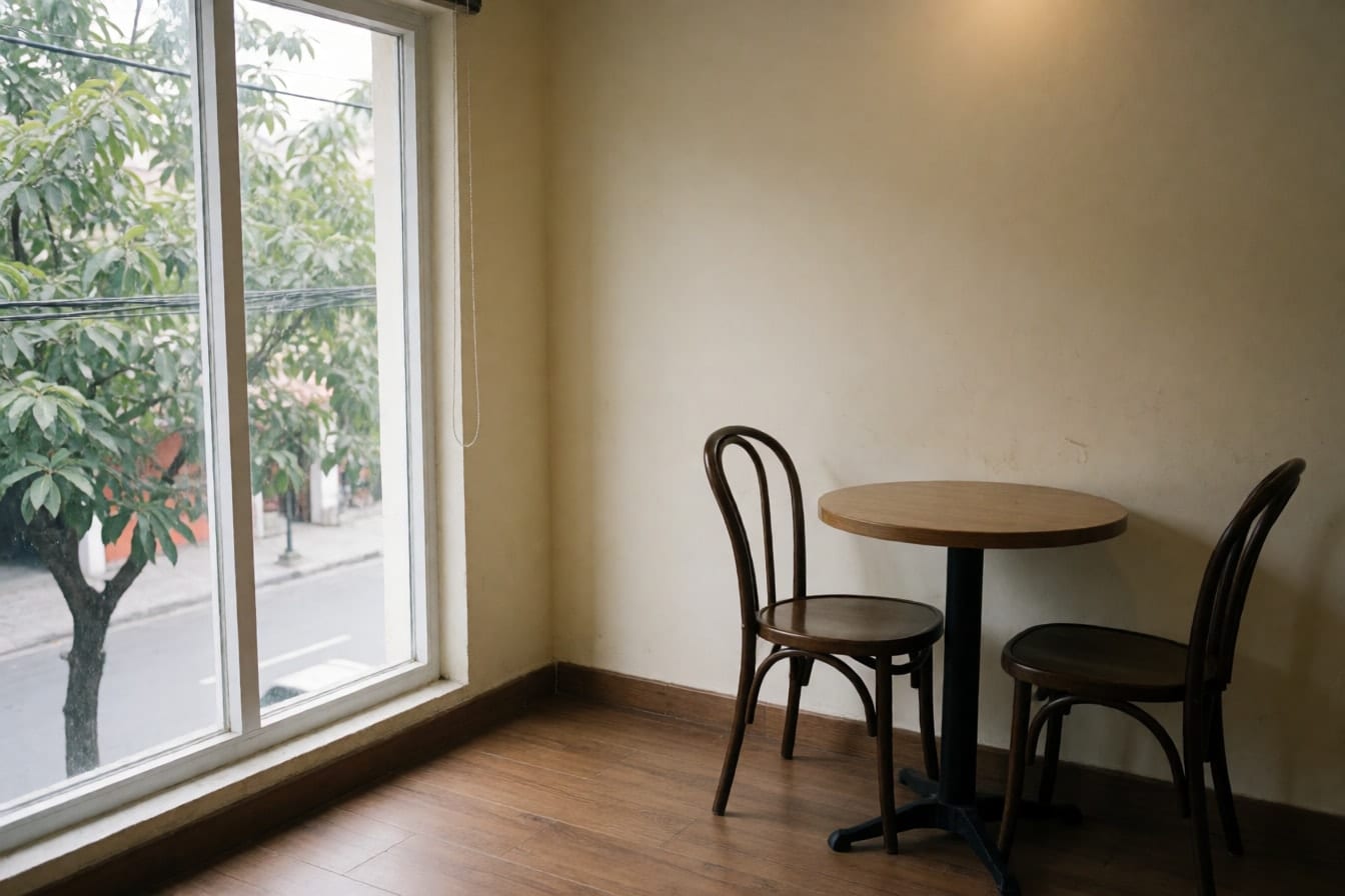 Góc trống ở tầng trên quán cà phê với cửa sổ lớn khung trắng nhìn ra hàng cây, tường kem, sàn gỗ và bàn tròn nhỏ hai ghế
