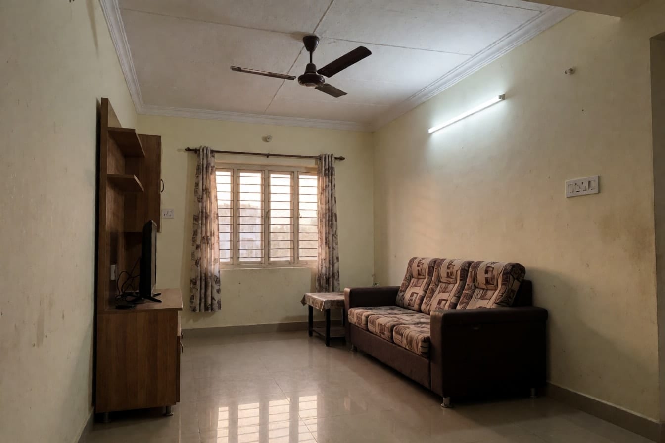 2BHK फ़्लैट का साधारण हॉल: सपाट सफ़ेद छत, बीच में पंखा, दीवार पर ट्यूबलाइट, कपड़े का सोफ़ा और ग्रिल वाली खिड़की