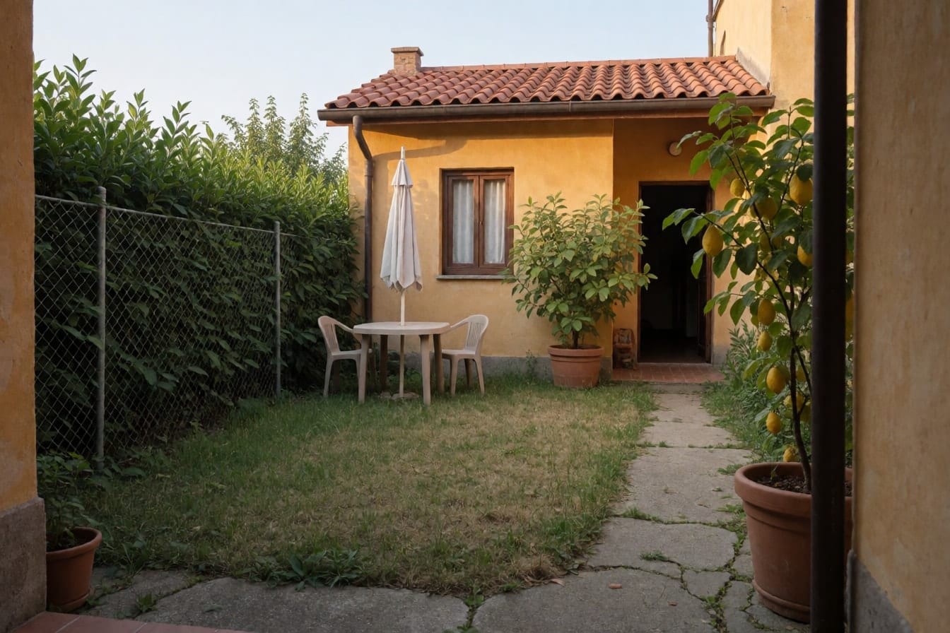 Giardino di una villetta con facciata ocra e tetto in coppi, prato a chiazze, vialetto di cemento crepato, siepe di lauro, tavolo con ombrellone e un limone in vaso