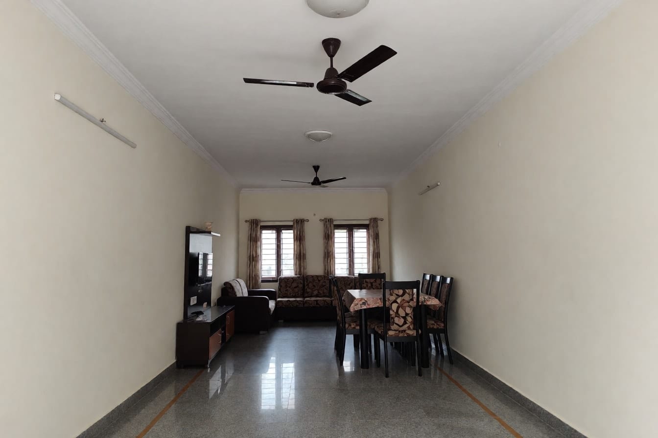 लंबा आयताकार हॉल: सपाट छत पर एक लाइन में दो पंखे, एक छोर पर L-आकार का सोफ़ा और दूसरे पर छह कुर्सियों वाली डाइनिंग टेबल