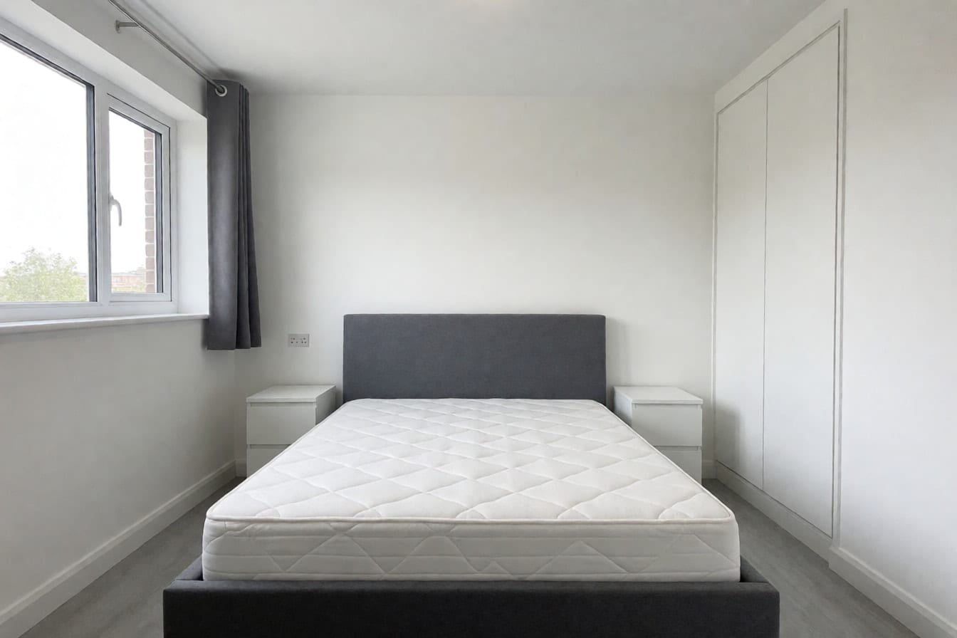 Schlafzimmer im Neubau mit grauem Polsterbett vor einer leeren weißen Wand, weißem Einbauschrank und hellgrauem Vinylboden