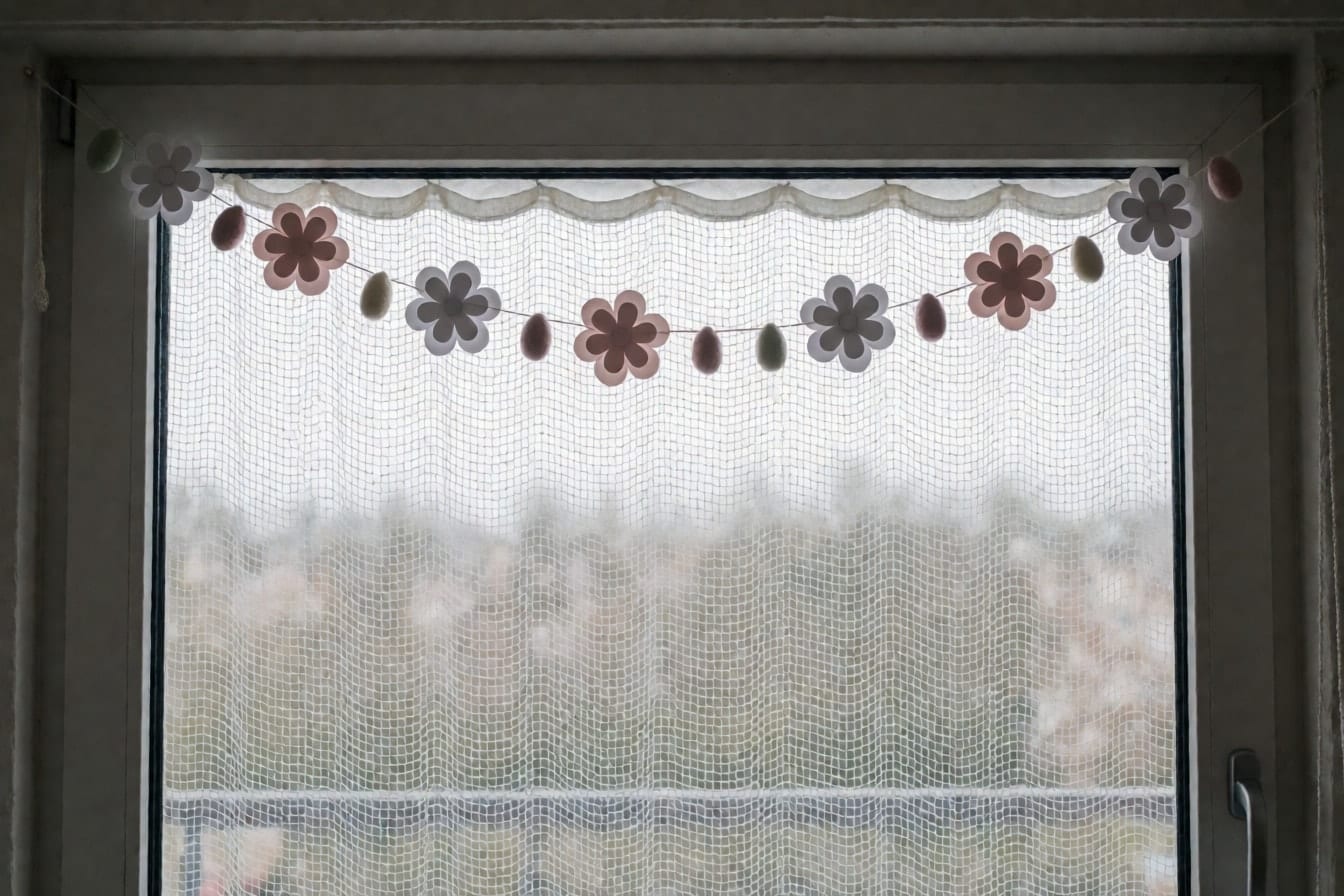 Girlande aus weißen und zartrosa Papierblumen und Filzeiern quer über einem Fenster