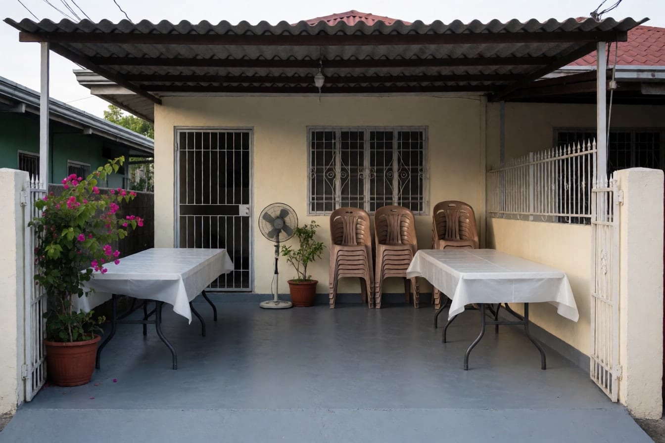 Carport ng bahay na may kulay-abong sementong sahig, cream na dingding, bakal na gate, dalawang mesang may puting plastik at nakapatong na monobloc