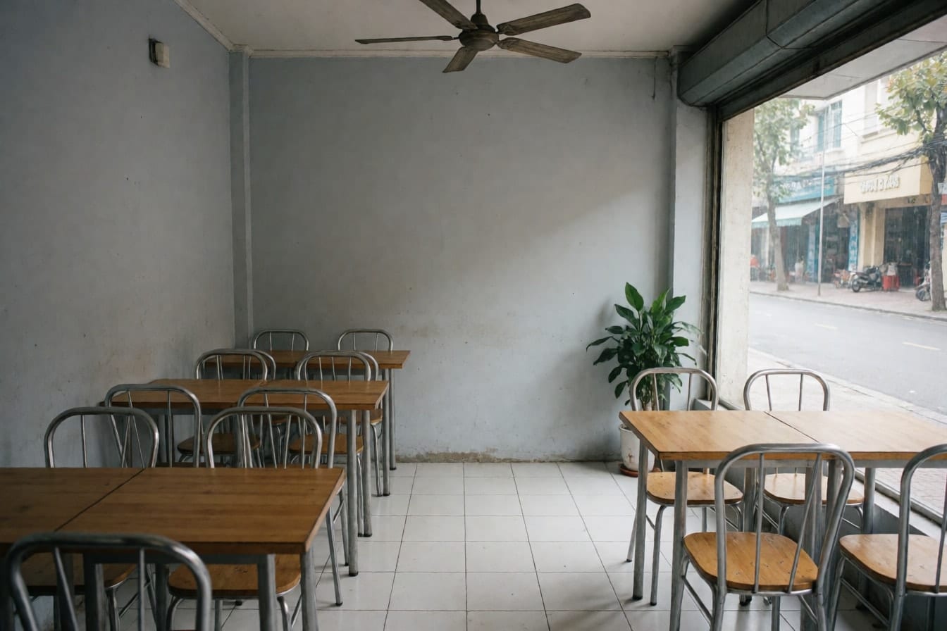 Khu ngồi tầng trệt của quán cà phê nhỏ với bàn gỗ vuông, ghế khung sắt, tường xám nhạt, sàn gạch trắng và quạt trần