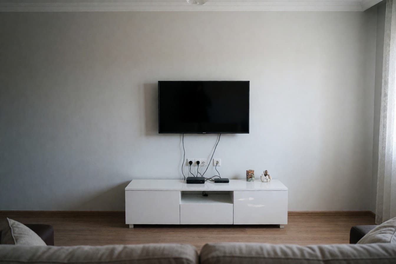 Açık gri boyalı duvarda asılı televizyon, altında parlak beyaz TV ünitesi ve görünen kablolar