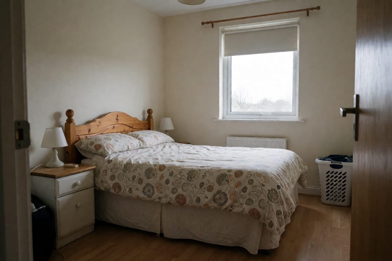 A plain rental bedroom with a pine headboard, a patterned duvet and a half-lowered roller shade
