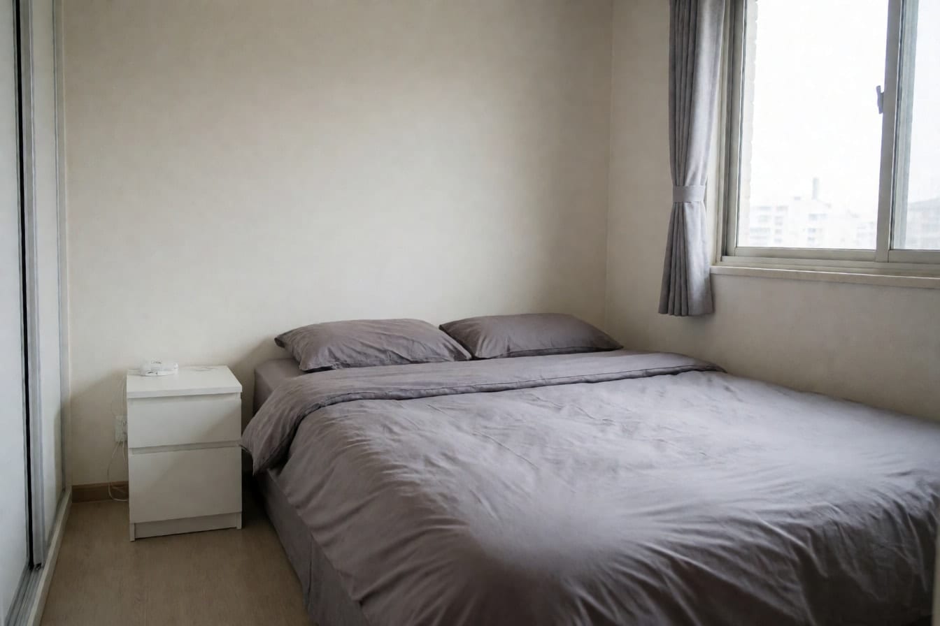 Lille soveværelse i lejlighed med dobbeltseng og grå dyne, hvidt sengebord, hvid garderobe med skydedøre og tyndt gråt gardin