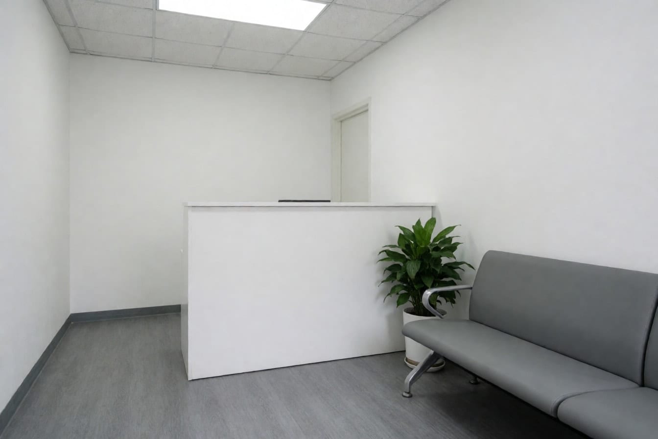 Khu lễ tân văn phòng nhỏ với quầy trắng trơn, ghế chờ xám, chậu cây, tường trắng, sàn vinyl xám và đèn panel trên trần