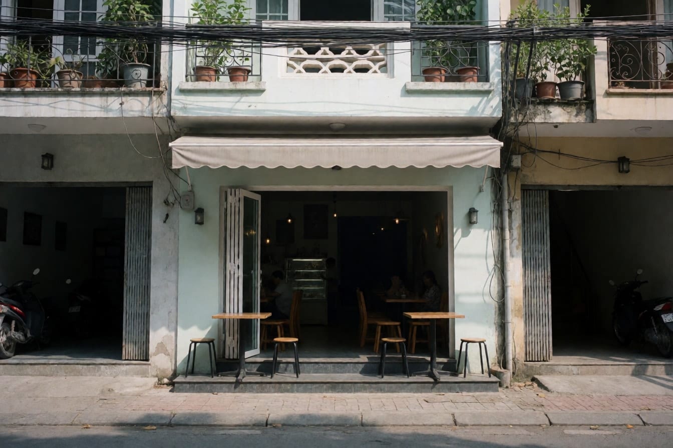 Mặt tiền quán cà phê ở tầng trệt nhà ống bốn tầng với cửa kính xếp mở, bàn ghế thấp dưới mái hiên và dây điện phía trước