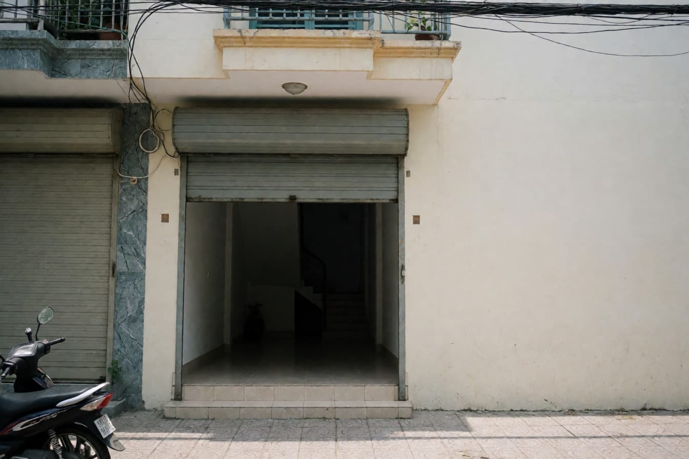 Tầng trệt nhà phố hẹp với cửa cuốn xám kéo lưng chừng, bậc thềm lát gạch, tường kem trơn và ban công nhỏ phía trên