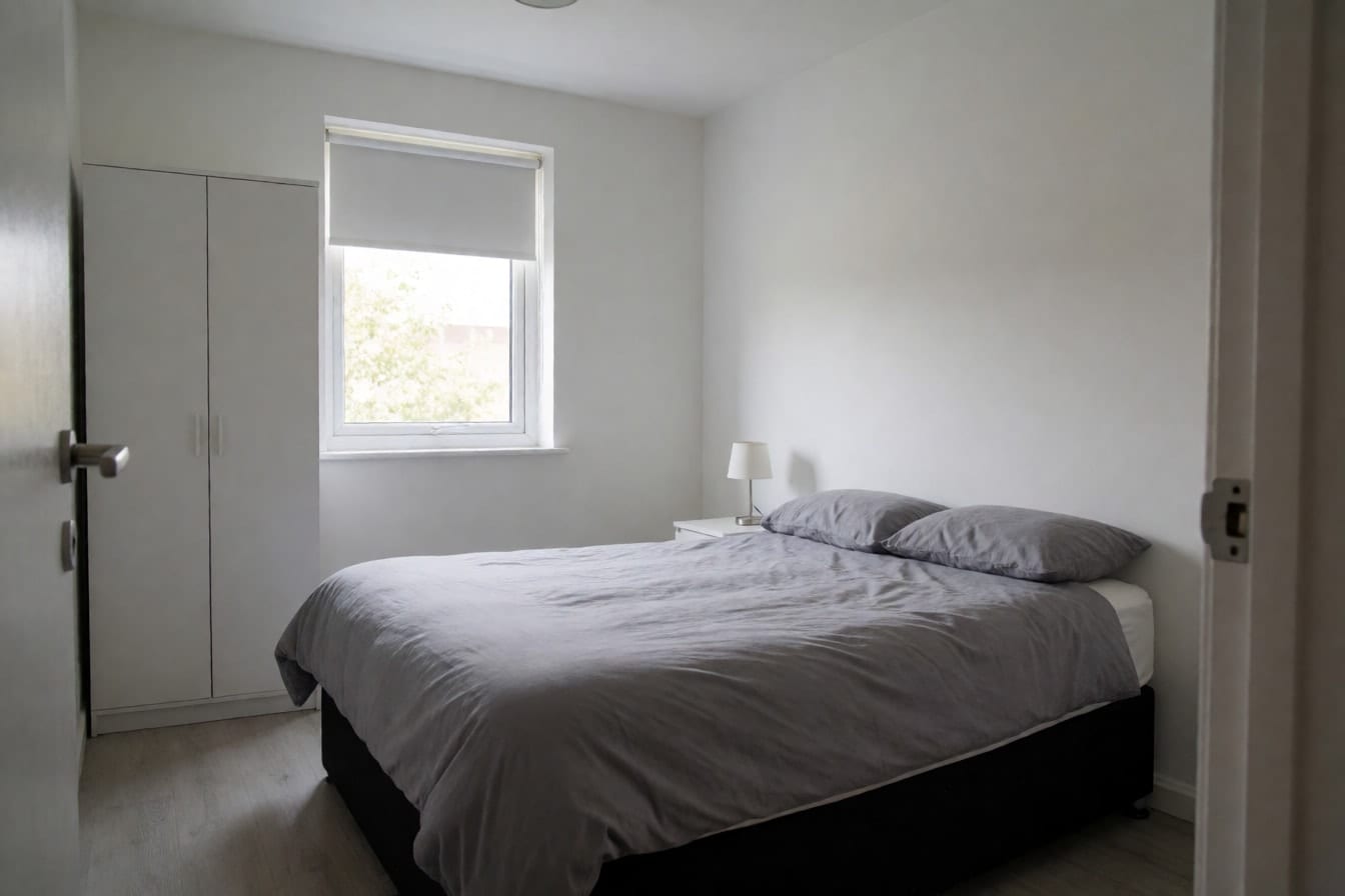 Dormitorio de piso de alquiler con cama de matrimonio, edredón gris, armario blanco de montar y paredes desnudas