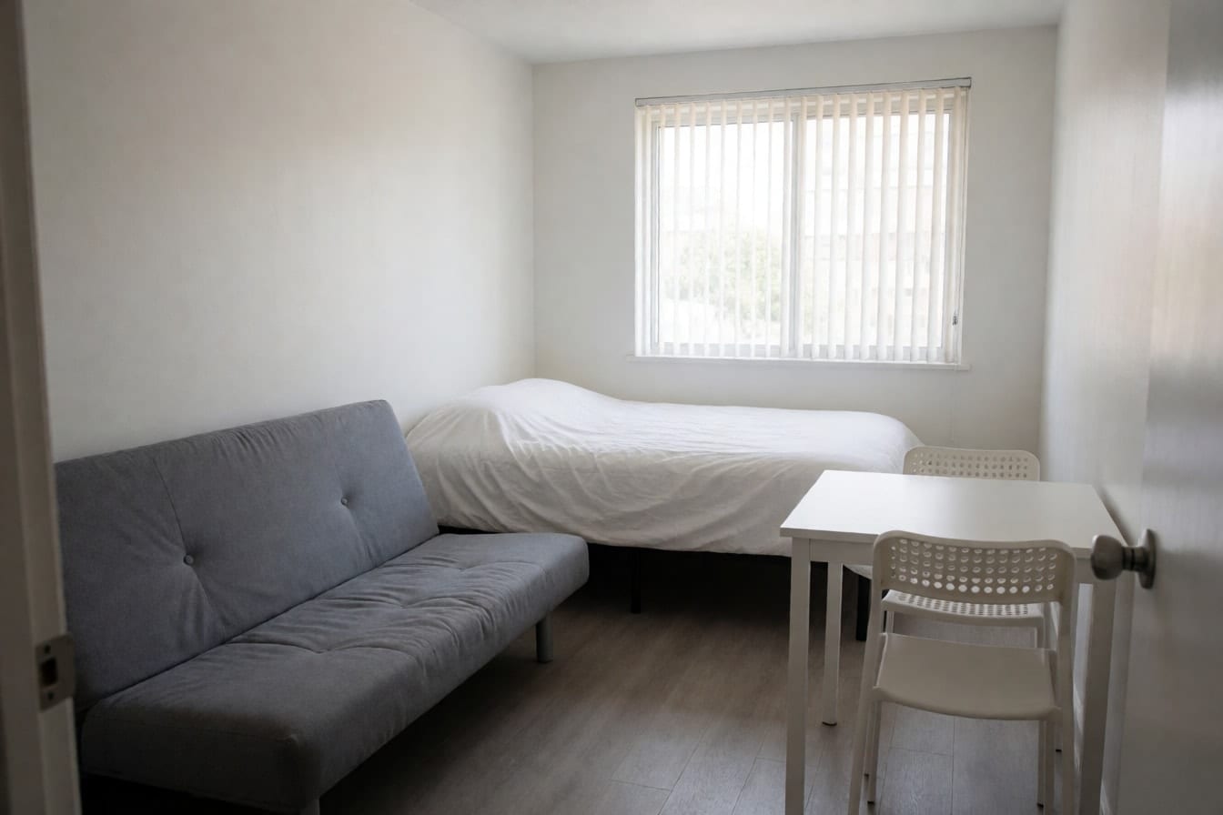 A bare rental studio with white walls, a grey futon, a double bed with a white duvet and vertical blinds