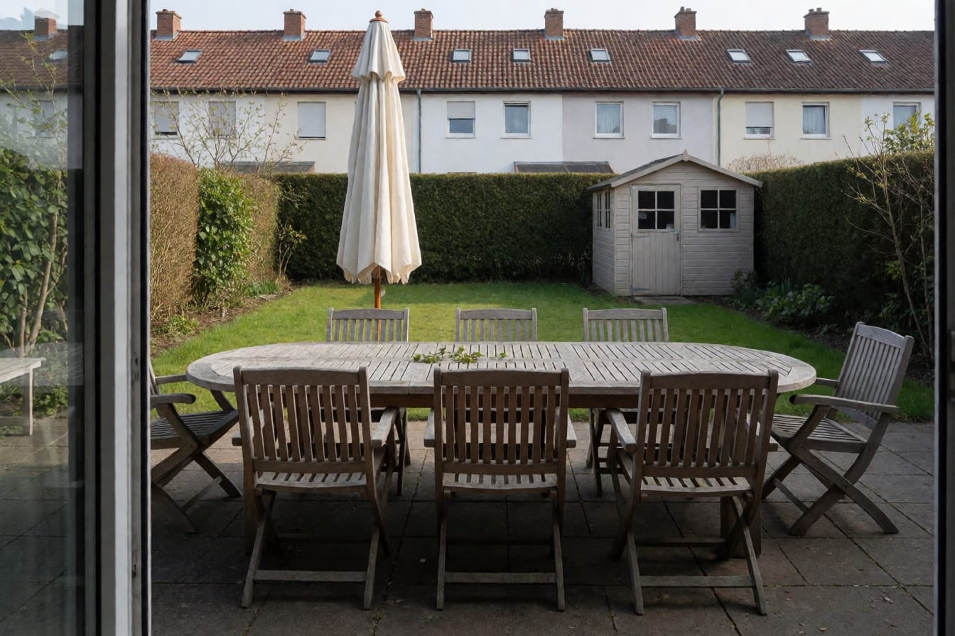 Gepflasterte Terrasse mit verwittertem Teaktisch, sechs Stühlen, geschlossenem Sonnenschirm, Rasen und Hecke