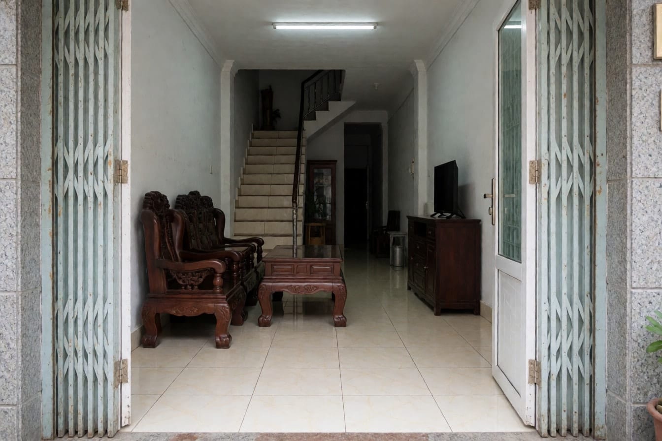 Phòng khách dài ở tầng trệt nhà ống, bộ sofa gỗ chạm, gạch men kem bóng, cầu thang ở cuối phòng và đèn tuýp trên trần