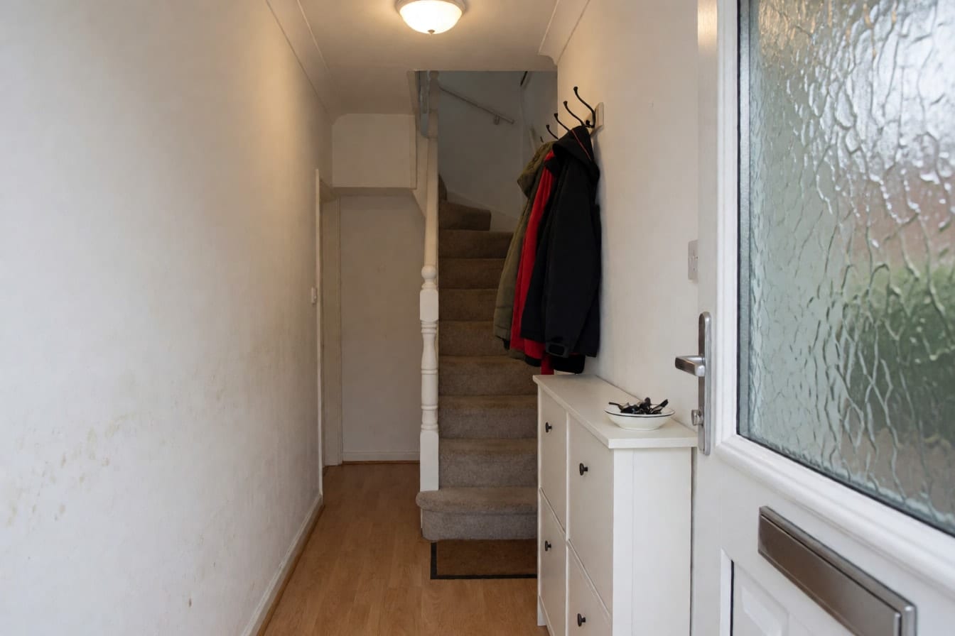 A family entrance hall with scuffed white walls, a white shoe cabinet, coat hooks and a white staircase banister