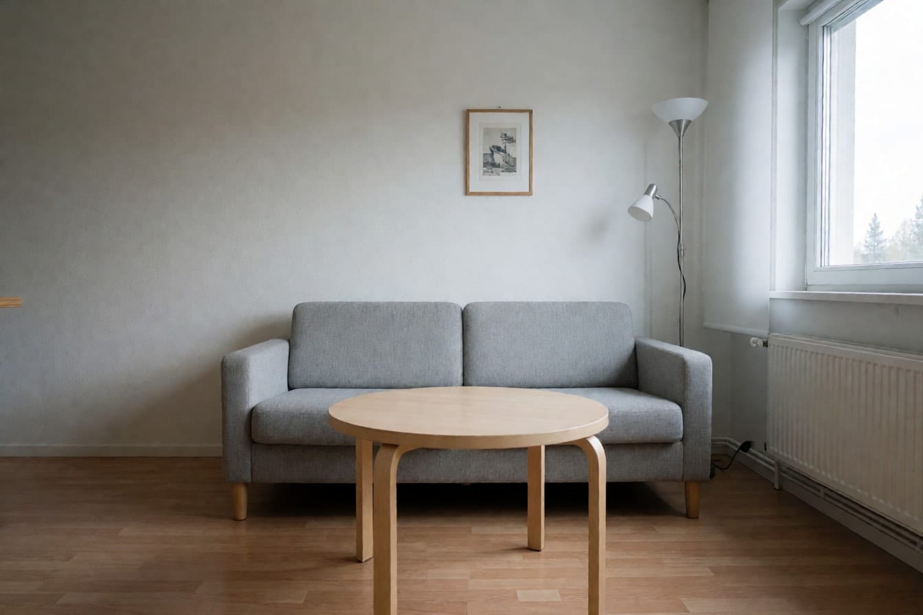 Pieni olohuone, jossa vaaleanharmaa sohva tasaista valkoista seinää vasten ja pyöreä koivuinen sohvapöytä